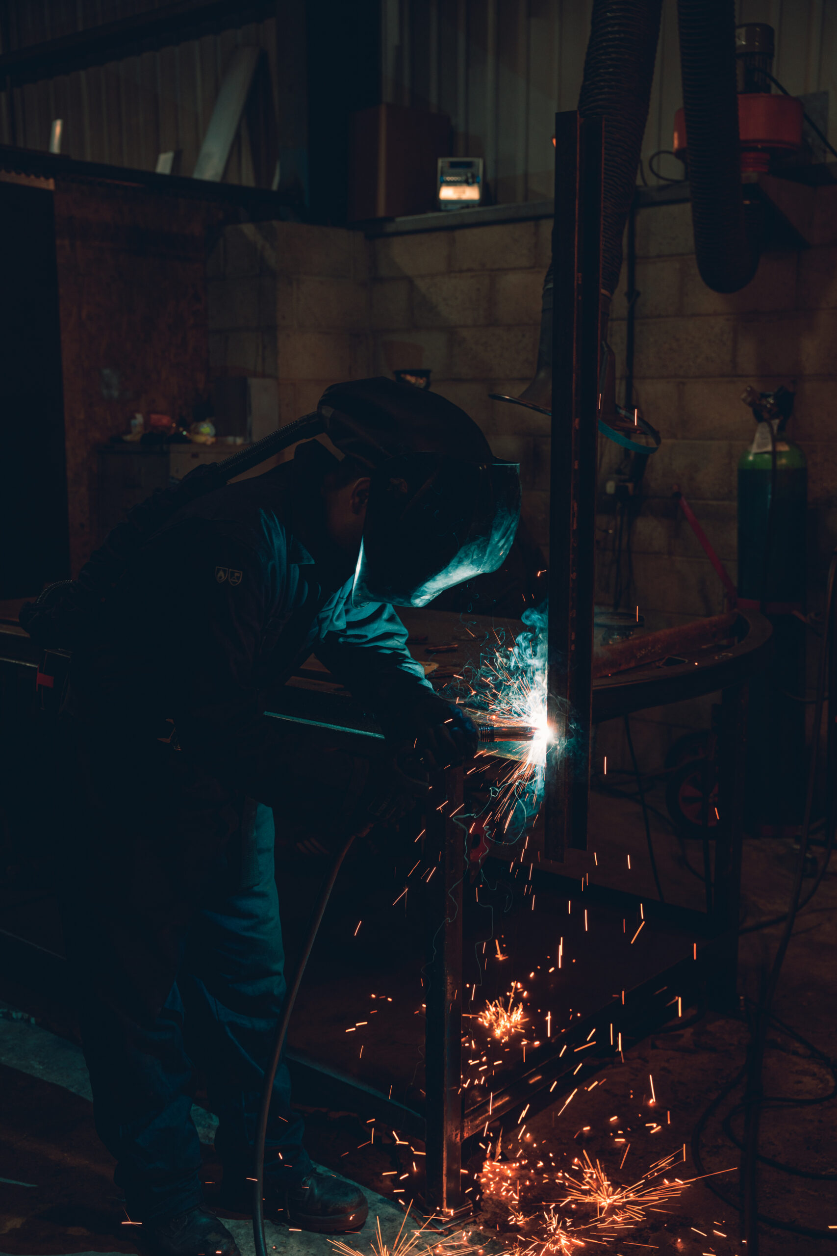A welder in a helmet joining a steel frame in the workshop, sparks falling to the floor